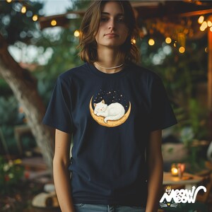 Op de afbeelding: Marineblauwe t-shirt met een afbeelding van een witte kat die slaapt op een maan, omringd door sterren. De tekst "Sip Meow" staat rechtsonder op het shirt. Het ontwerp is gecentreerd op het shirt.