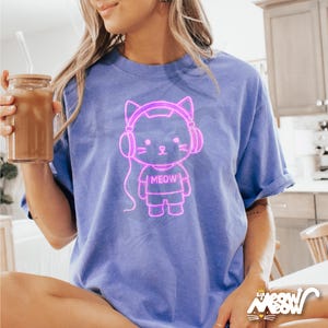 Op de afbeelding: Een paars T-shirt met een neonroze omtrek van een kat met een koptelefoon en het woord "MEOW" op zijn shirt.