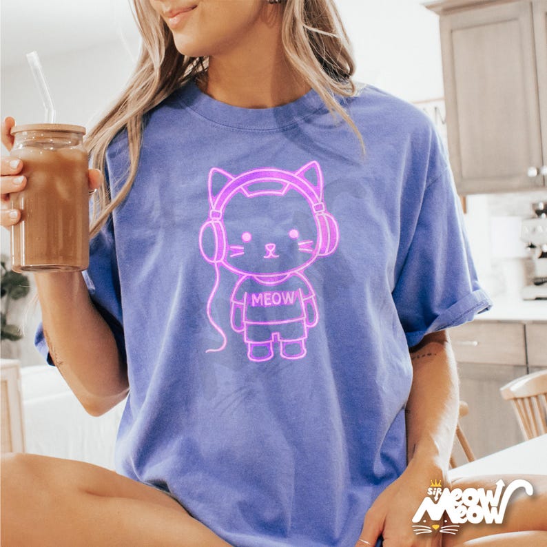 Op de afbeelding: Een paars T-shirt met een neonroze omtrek van een kat met een koptelefoon en het woord "MEOW" op zijn shirt.