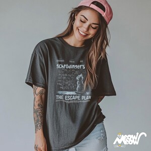 May include: A woman wearing a black t-shirt with a graphic of a cat and the text "Schr&ouml;dinger's The Escape Plan". The shirt has a distressed look and the cat is looking up. The woman is wearing a pink baseball cap and has long brown hair.