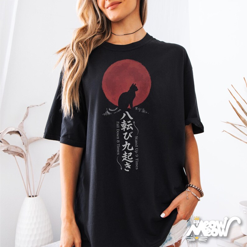 Op de afbeelding: Zwart T-shirt met een katten silhouet voor een rode cirkel, met Japanse tekst eronder. De tekst luidt "Stand Up 9 Times, Fall Down 8 Times". Het shirt heeft korte mouwen en een ronde hals.
