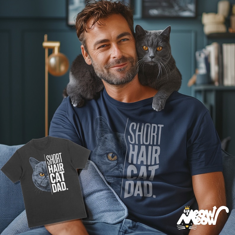 May include: A man wearing a navy blue t-shirt with the text "Short Hair Cat Dad." A gray cat is sitting on his shoulder. The man is also wearing a gray t-shirt with the same text and a cat image.