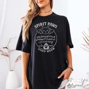 May include: Black t-shirt with a white graphic featuring a cat with bat wings, a planchette, and the words "Spirit Paws." The design includes the alphabet, numbers, and the words "Purr," "Hiss," "Good," and "Meow."