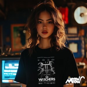 Peut inclure: Une femme portant un t-shirt noir avec un imprim&eacute; graphique blanc d'une affiche de film. L'affiche pr&eacute;sente le texte "The Watchers" et un graphique d'une voie ferr&eacute;e avec des chats qui marchent dessus. Le texte "Double Split Productions Present" est en haut de l'affiche.
