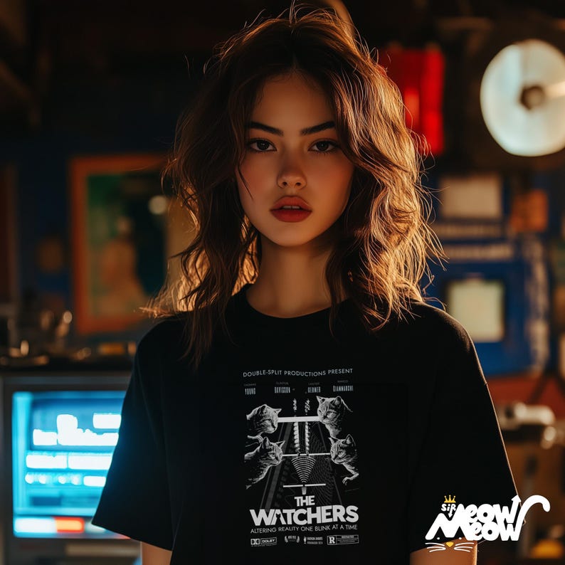 Peut inclure: Une femme portant un t-shirt noir avec un imprim&eacute; graphique blanc d'une affiche de film. L'affiche pr&eacute;sente le texte "The Watchers" et un graphique d'une voie ferr&eacute;e avec des chats qui marchent dessus. Le texte "Double Split Productions Present" est en haut de l'affiche.