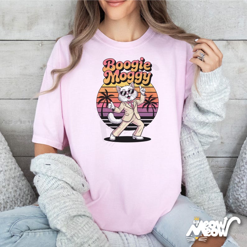 May include: Light pink t-shirt featuring a cartoon cat in a suit and sunglasses, with the text "Boogie Moggy" above it. The cat is dancing in front of a sunset graphic with palm trees. The shirt has short sleeves.
