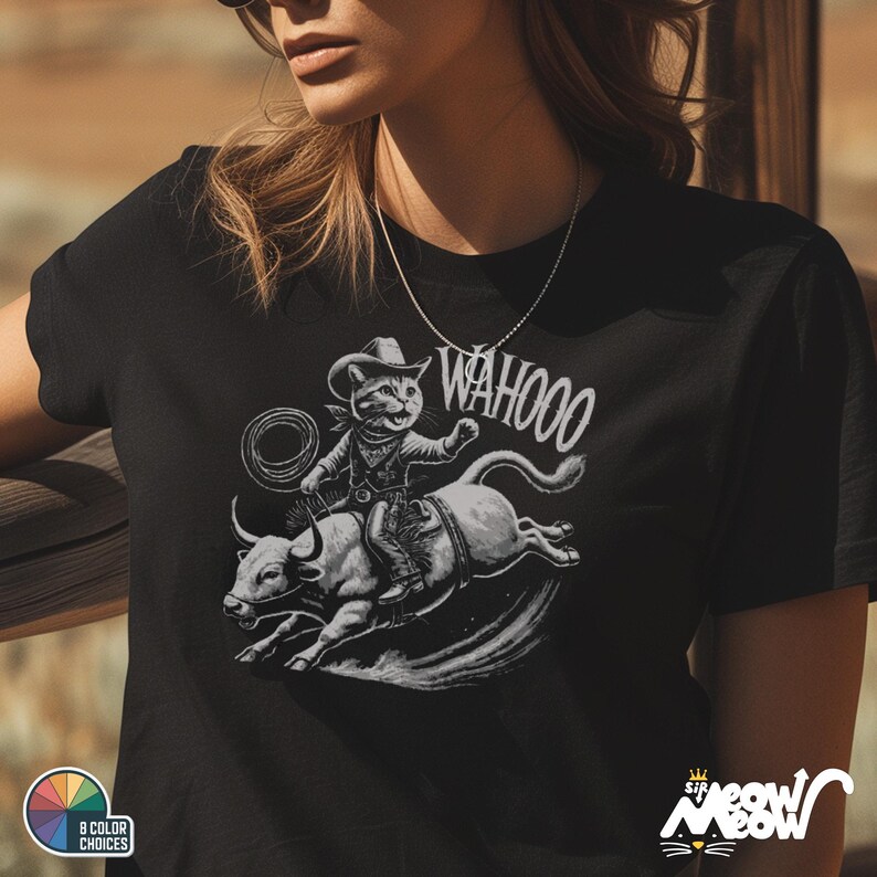 May include: A black t-shirt with a white graphic of a cat wearing a cowboy hat and riding a bull. The text "WAHOO!" is printed above the cat.