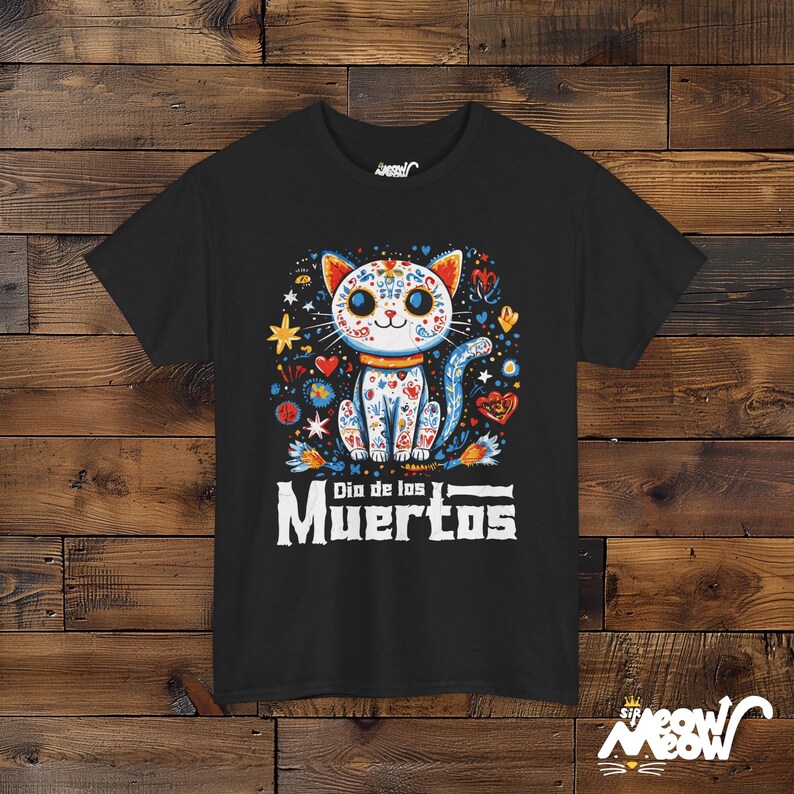 May include: Black t-shirt featuring a colorful cat design with floral and heart patterns, and the text "Dia de los Muertos". The cat has a white face and body with blue, orange, and red accents. The shirt has a crew neck.