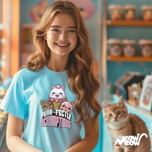 May include: A light blue t-shirt with a graphic of three cartoon cats in ice cream cones. The text "Purr-fectly Scrumptious" is printed below the cats. A ginger cat is sitting in the foreground.