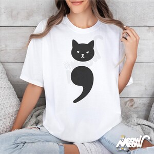 May include: A white t-shirt with a black cat graphic and a black semicolon. The cat is looking to the left and has a grumpy expression.