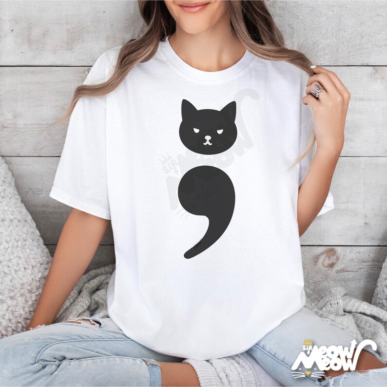 May include: A white t-shirt with a black cat graphic and a black semicolon. The cat is looking to the left and has a grumpy expression.