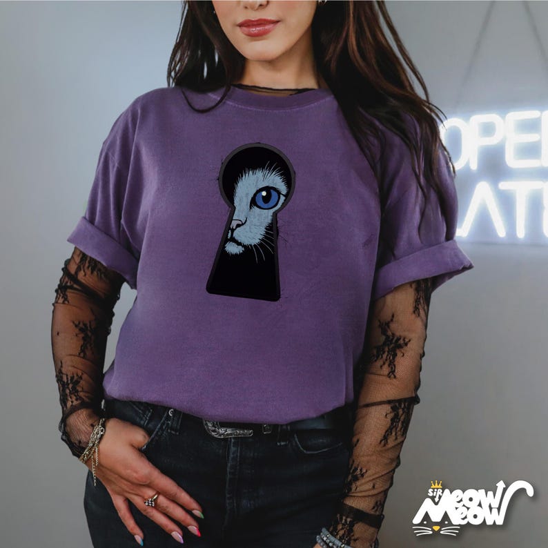 Pu&ograve; includere: Una maglietta viola con una grafica di un viso di gatto che guarda attraverso un buco della serratura.