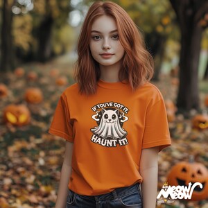 以下が含まれることがあります： 「IF YOU'VE GOT IT, HAUNT IT!」の文字と、漫画のゴーストキャットのデザインが施されたオレンジ色のTシャツ。Tシャツを着ている人は赤毛です。背景にはカボチャと紅葉が見えます。