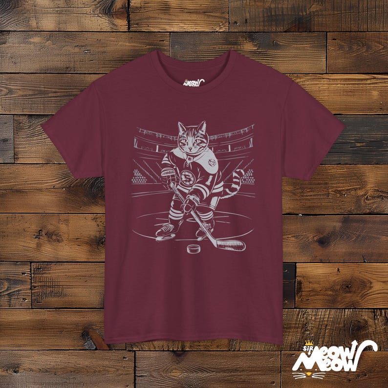 Op de afbeelding: Bordeauxrood T-shirt met een witte illustratie van een kat die hockey speelt. De kat draagt een hockeytrui en houdt een hockeystick vast. De tekst "Sir Meow" is gedrukt in de rechterbenedenhoek van het shirt.