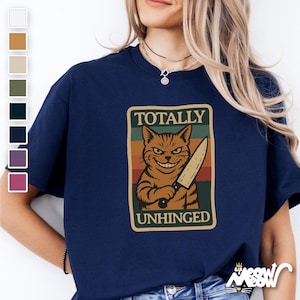 May include: A navy blue t-shirt with a vintage-style graphic of a cat holding a knife. The graphic says "Totally Unhinged".