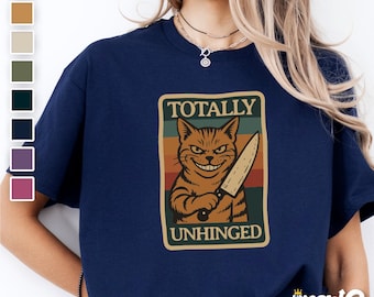 May include: A navy blue t-shirt with a vintage-style graphic of a cat holding a knife. The graphic says "Totally Unhinged".