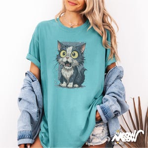 May include: Teal t-shirt with a cartoon cat illustration. The cat has large yellow eyes and an open mouth, appearing surprised. It's in grey and white, with water droplets around it. The shirt has rolled sleeves and a loose fit. The word "Meow" is in the bottom right corner.