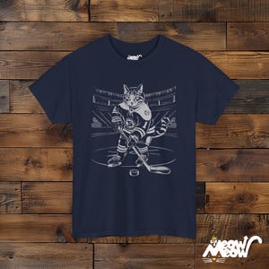 Op de afbeelding: Een marineblauw t-shirt met een afbeelding van een kat die hockey speelt. De kat draagt een hockeytrui en houdt een hockeystick vast. De afbeelding is in wit en grijs.