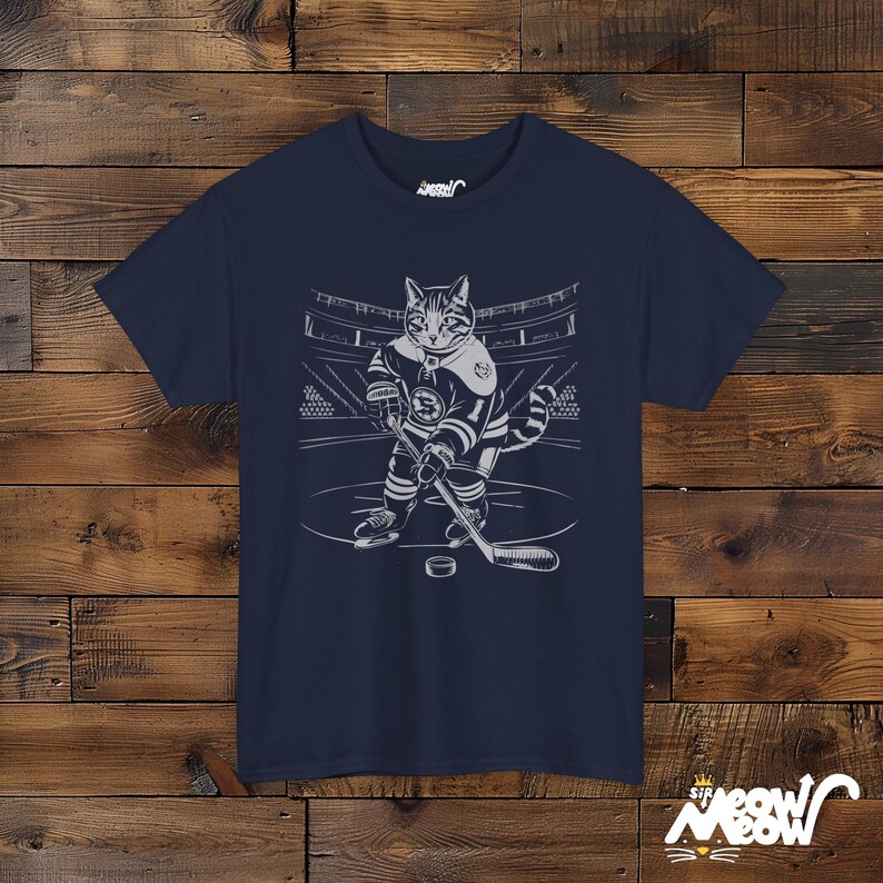 Op de afbeelding: Een marineblauw t-shirt met een afbeelding van een kat die hockey speelt. De kat draagt een hockeytrui en houdt een hockeystick vast. De afbeelding is in wit en grijs.