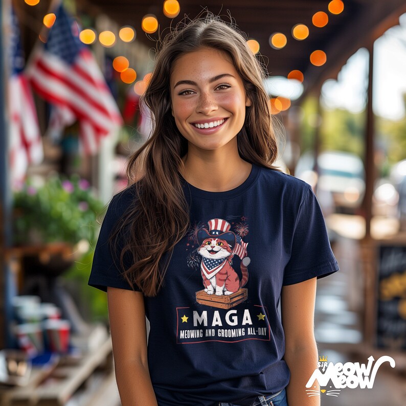 May include: A navy blue t-shirt with a cartoon cat wearing a red, white, and blue hat and holding a firework. The cat is standing on a wooden crate. The text on the shirt reads "Maga, Meowing and Grooming All-Day".
