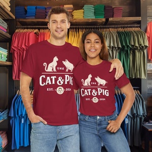 K&ouml;nnte beinhalten: Zwei Personen tragen passende weinrote T-Shirts mit dem Text "The Cat & Pig" und dem Gr&uuml;ndungsjahr "2024". Die Shirts zeigen eine Katze und ein Schwein. Sie stehen in einem Bekleidungsgesch&auml;ft.