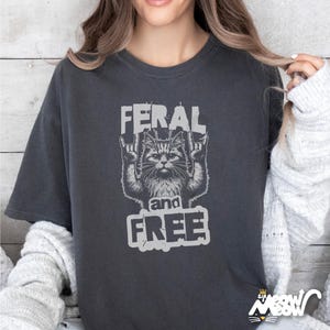 Puede incluir: Una camiseta gris oscuro con un gr&aacute;fico de un gato con un gesto de rock and roll. El texto "Feral and Free" est&aacute; impreso debajo del gato.