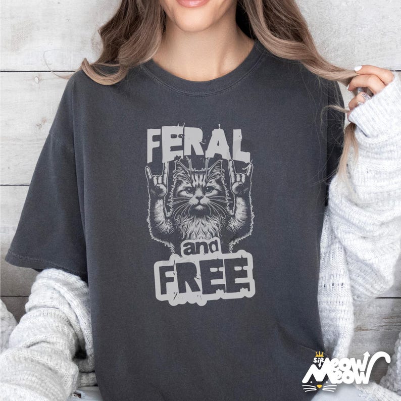 Puede incluir: Una camiseta gris oscuro con un gr&aacute;fico de un gato con un gesto de rock and roll. El texto "Feral and Free" est&aacute; impreso debajo del gato.