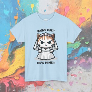 May include: Light blue t-shirt with a cartoon illustration of a cat wearing a white wedding dress and a flower crown. The text "PAWS OFF!! HE'S MINE!!" is printed on the shirt.