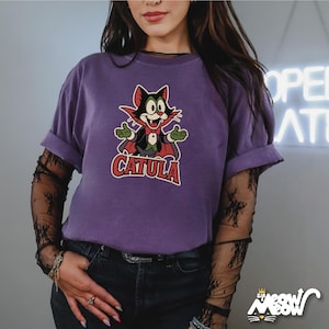 May include: A purple t-shirt featuring a cartoon cat character with the text "CATULA". The cat is dressed in a vampire-like outfit with a red cape and bow tie. The shirt has rolled-up sleeves.