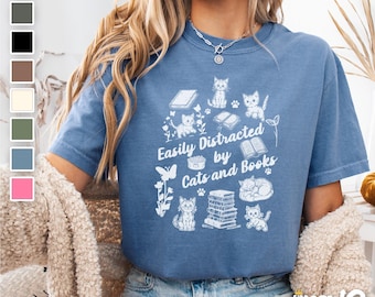 May include: A blue t-shirt with a white graphic design featuring cats, books, and the text "Easily Distracted by Cats and Books".