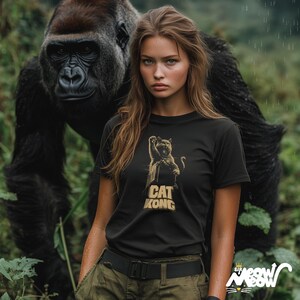 Może przedstawiać: Kobieta w czarnym t-shircie z złotym nadrukiem kota z napisem "CAT KONG". Stoi przed gorylem w dżungli.
