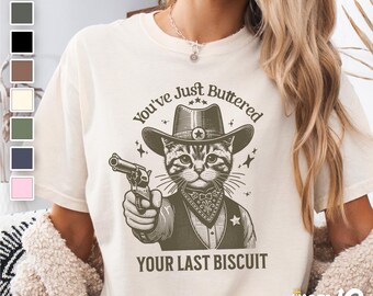 May include: A cream-colored t-shirt with a cartoon cat wearing a cowboy hat and holding a gun. The text on the shirt reads "You've Just Buttered Your Last Biscuit".