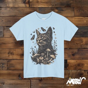 Vintage style t-shirt print of a cat in the woodlands surrounded by wildflowers, mushrooms and butterflies. light blue tee.
