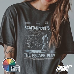 May include: A dark gray t-shirt with a white graphic of a cat looking up at a chalkboard with equations and the text "Schrödinger's The Escape Plan".