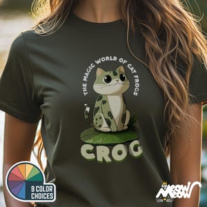 May include: A dark green t-shirt with a cartoon cat wearing a green frog costume. The text on the shirt reads "The Magic World of Cat Frogs, Croc".