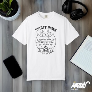 May include: White t-shirt with a black graphic featuring a cat-shaped planchette, a bat, and the words "Spirit Paws," "Good Meow," and the alphabet. The design is reminiscent of a Ouija board. The shirt is displayed on a wooden surface.