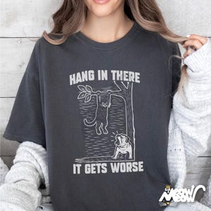 May include: Dark gray t-shirt with a white graphic of a cat hanging from a tree branch and a dog looking up. The text "HANG IN THERE" is above the cat, and "IT GETS WORSE" is below. The shirt has a relaxed fit.