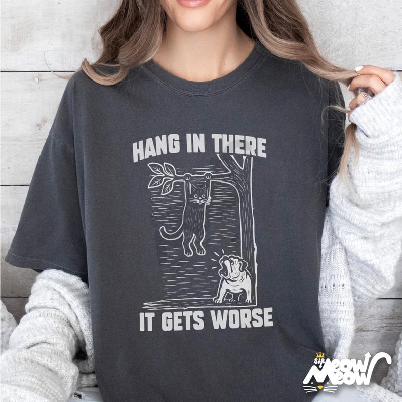 May include: Dark gray t-shirt with a white graphic of a cat hanging from a tree branch and a dog looking up. The text "HANG IN THERE" is above the cat, and "IT GETS WORSE" is below. The shirt has a relaxed fit.