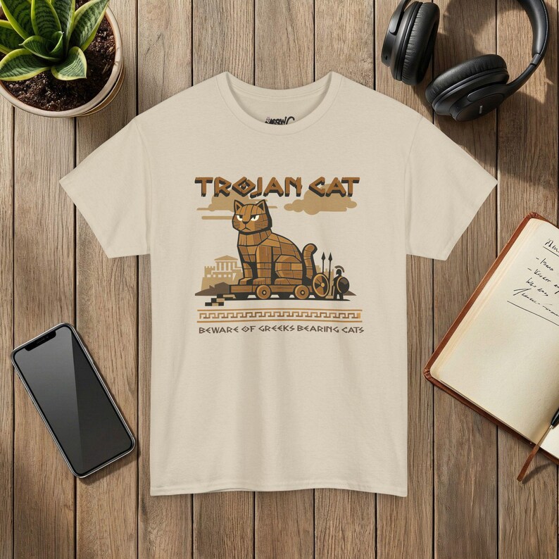 May include: A beige short-sleeve t-shirt featuring a graphic of a wooden cat on wheels. The text reads TROJAN CAT and BEWARE OF GREEKS BEARING CATS. The design includes a small warrior figure with a spear and shield, set against a simple landscape.