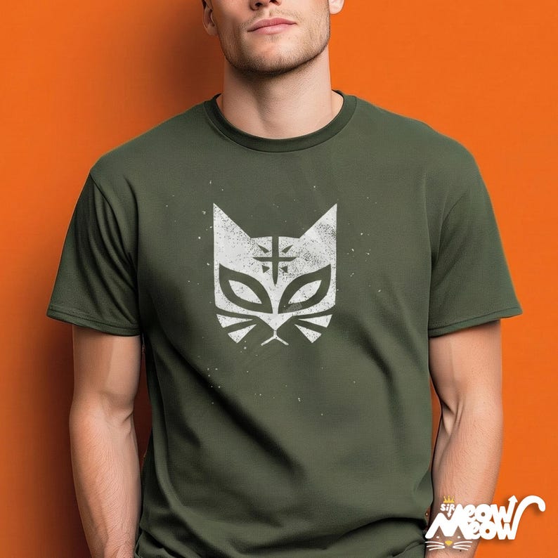 May include: Olive green t-shirt featuring a white cat face design with a cross symbol on the forehead. The graphic has a distressed, vintage look. The shirt is short-sleeved and has a crew neck. The background is orange.