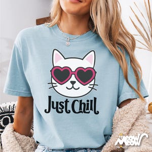 Pu&ograve; includere: T-shirt azzurra con la faccia di un gatto bianco con occhiali da sole rosa a forma di cuore. Il testo "Just Chill" &egrave; stampato sotto il disegno del gatto. La maglietta ha maniche corte e girocollo.