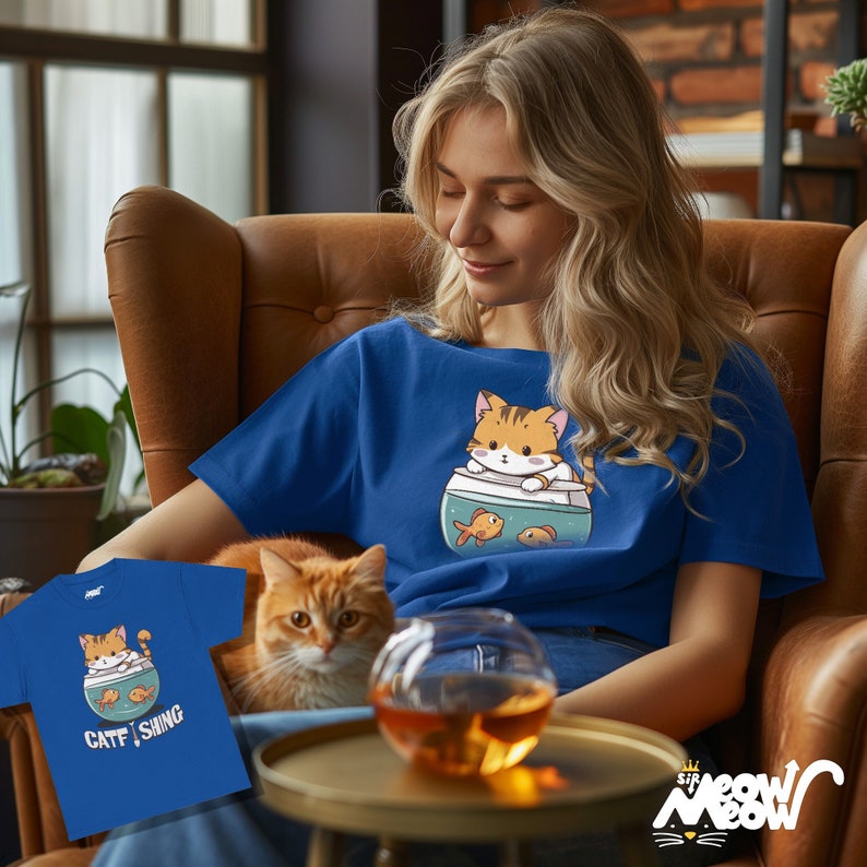 May include: Royal blue t-shirt with a cartoon cat in a fishbowl and two goldfish. The shirt has the text "CATFISHING" on a smaller version of the shirt. The cat is orange and white. The shirt is worn by a person.