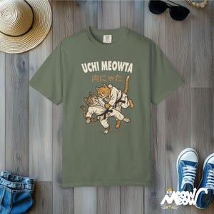 K&ouml;nnte beinhalten: Ein olivgr&uuml;nes T-Shirt mit einer Grafik von zwei Katzen in wei&szlig;en Kampfkunstanz&uuml;gen mit schwarzen G&uuml;rteln, die einen Judowurf ausf&uuml;hren. Der Text UCHI MEOWTA und japanische Schriftzeichen sind in cremefarbener Schrift &uuml;ber den Katzen gedruckt.
