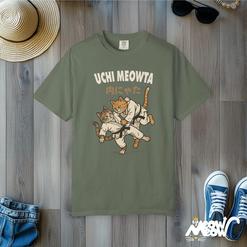 K&ouml;nnte beinhalten: Ein olivgr&uuml;nes T-Shirt mit einer Grafik von zwei Katzen in wei&szlig;en Kampfkunstanz&uuml;gen mit schwarzen G&uuml;rteln, die einen Judowurf ausf&uuml;hren. Der Text UCHI MEOWTA und japanische Schriftzeichen sind in cremefarbener Schrift &uuml;ber den Katzen gedruckt.