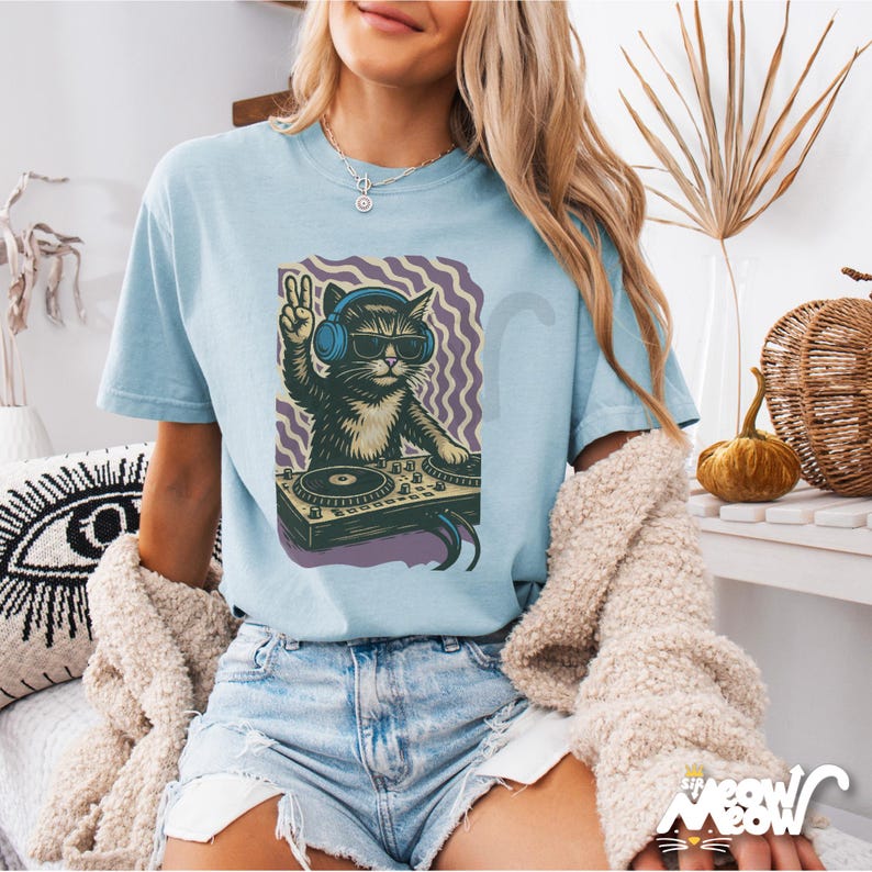May include: Light blue t-shirt featuring a graphic of a cat DJ wearing sunglasses and headphones, making a peace sign. The cat is behind a turntable with a purple and white wavy background. The text "The Meow Meow" is at the bottom right.