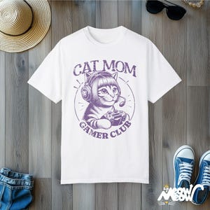 Cat Parent Gamer Club T-Shirts. The tee shows a cat holding a gaming controller. Above the image is the text "Cat Mom" and below the image is the text "Gamer Club".
The t-shirt shown is in the color "White" and is also available in a "Cat Dad" version.