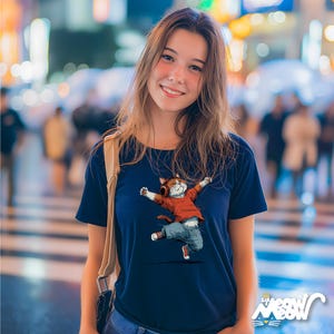 May include: Navy blue t-shirt featuring a dancing cat wearing headphones, a red shirt, and blue jeans. The cat has its arms outstretched and is wearing red sneakers. The shirt has the text "sip meow" in the bottom right corner.