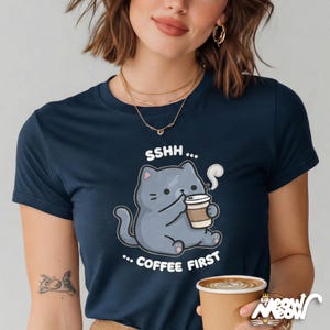 May include: Navy blue t-shirt featuring a cartoon cat holding a coffee cup. The cat is gray with black details, and the shirt has the text "SSHH... COFFEE FIRST". The shirt is a crew neck.