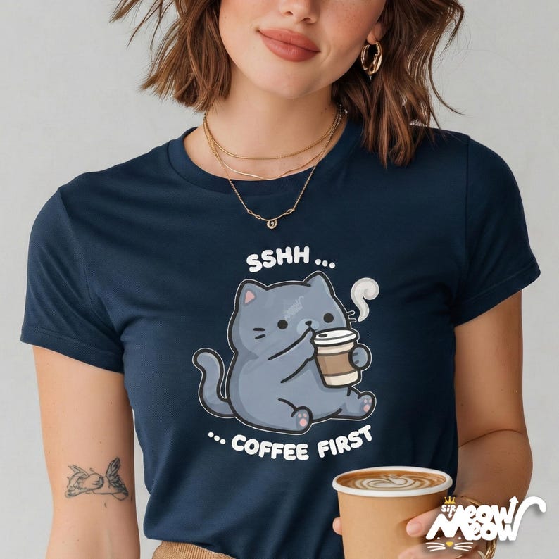 May include: Navy blue t-shirt featuring a cartoon cat holding a coffee cup. The cat is gray with black details, and the shirt has the text "SSHH... COFFEE FIRST". The shirt is a crew neck.