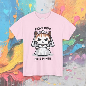 May include: Pink t-shirt with a cartoon illustration of a cat wearing a white wedding dress and a flower crown. The text "PAWS OFF!! HE'S MINE!!" is printed on the shirt.